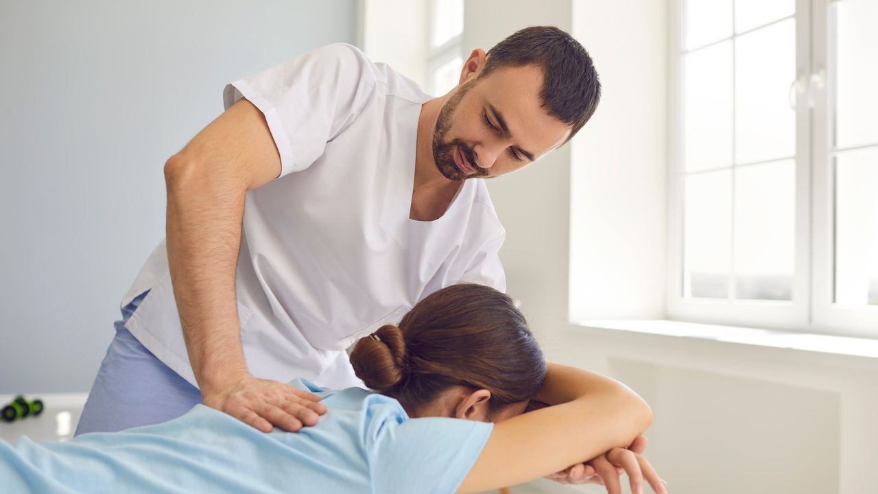 Therapeut mobilisiert mit beiden Händen die Schulter einer bäuchlings liegenden Patientin
